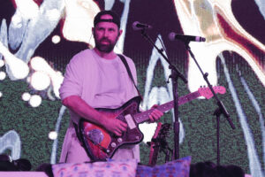 Bearded guitarist wearing a backward cap and white shirt playing a sunburst electric guitar on a stage with microphones behind him.