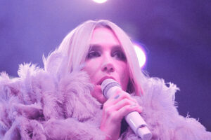 Close-up of a blonde singer on stage, wearing a fluffy feather boa and singing into a microphone under purple lights.