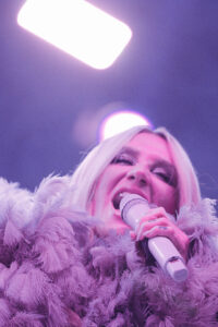 Female singer on stage, holding a microphone, wearing a purple feathered coat under purple lighting.