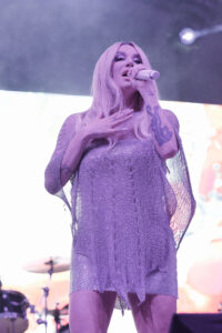 Female singer with long blonde hair in a shimmering purple mesh dress, performing on stage.