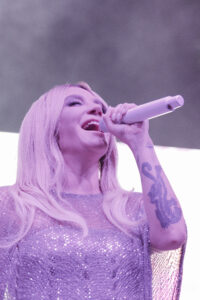 Female singer on stage, holding a microphone and singing with closed eyes, wearing a sparkly purple outfit.