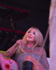 Smiling blonde female performer in a glittery silver outfit holds a microphone on stage with pink lighting in the background.