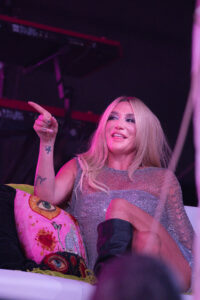 Smiling woman with long blonde hair in a glittery silver dress, pointing upward on a stage with pink lighting.