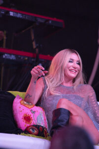 Smiling woman with long blonde hair in a glittery silver dress on stage, seated with a pink patterned pillow beside her
