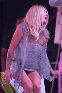 Female singer on stage in a glittering silver outfit, holding a microphone with pink stage lighting and tattoos visible on her arm.