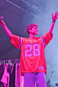 Concert performer on stage in an orange jersey with the number 28, microphone in hand and arms raised under pink lighting.