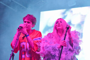 Two performers sing into microphones on stage, bathed in pink and blue light, wearing bold colorful outfits.