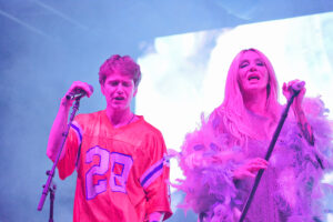 Two singers perform on stage under blue and pink lights, each holding a microphone.