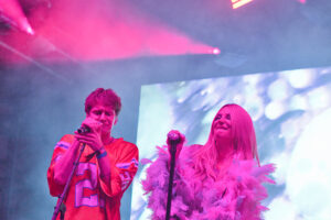 Two performers sing into microphones on a brightly lit stage with pink and purple lighting behind them.