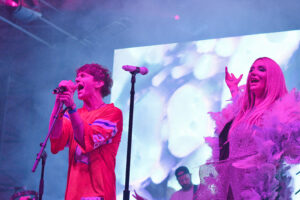 Two bold performers sing into microphones on a colorful stage with purple lighting and a bright backdrop.
