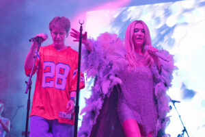 Two performers sing into microphones on a vibrant, purple-lit stage; man in an orange jersey with number 28, woman in a glittery dress with a pink feather boa nearby.