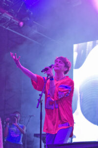 Male singer on stage, pink outfit, holding a microphone and reaching out to the crowd under purple lights.