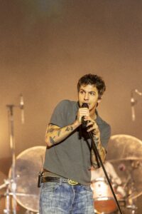 Male singer on stage holding a microphone, performing with tattoos visible on his arms.