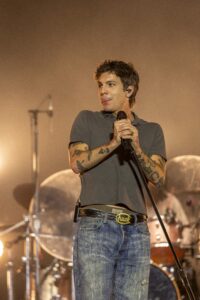 Male singer with tattoos gripping a microphone on stage, wearing a gray polo and jeans amid warm stage lights and a drummer in the background.