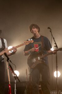 Male guitarist on stage with a brown electric guitar, wearing a dark Nike shirt, under warm concert lighting.