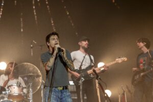 Frontman singing into a microphone on stage, with band members playing guitars and drums in the background under warm stage lights.