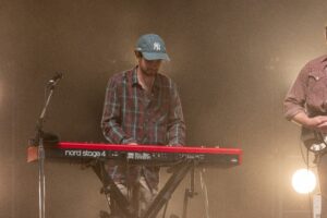 Keyboardist wearing a plaid shirt and blue cap plays a red Nord Stage 4 keyboard on a dim stage, with a microphone stand nearby.