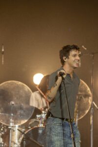 Male singer on stage, holding a microphone and smiling, with drums and bright stage lights in the background.