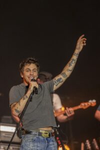 Male singer on stage raises his tattooed arm while singing into a microphone on stage lights behind him