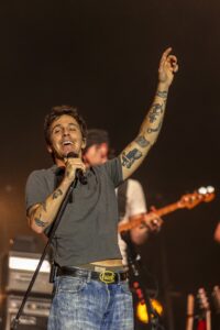 Male singer on stage, microphone in hand, arm raised in a fist as he performs; visible tattoos on his forearm.