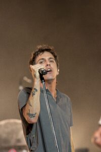 Male singer wearing a gray shirt, holding a microphone on stage, tattoos visible on his arm
