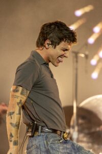Male musician on stage, wearing a gray shirt and jeans, with a wired in-ear monitor and belt pack, sticking out his tongue during a performance.