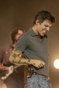 Male musician on stage, smiling with eyes closed, wearing a gray polo and jeans with tattooed arms.