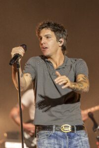 Male singer on stage in a gray polo shirt, jeans, and visible arm tattoos, singing into a handheld microphone.