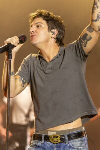 Male singer on stage, holding a microphone and raising one arm, wearing a gray polo and jeans with a decorative belt buckle, visible tattoos on arms