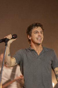 Male singer on stage holding a microphone, smiling, wearing a gray polo shirt with visible tattoos on his arm.