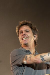Male performer on stage smiling, with short brown hair and visible tattoos on his right forearm, wearing a gray shirt