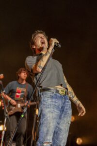 Singer passionately performs on stage, gripping a microphone with tattoos visible on his arm, wearing a gray shirt and ripped jeans.