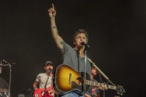 Male guitarist performs on stage, raising his right arm with index finger pointing up while singing into a microphone.