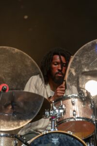 Drummer with dreadlocks playing a sparkling drum kit on stage, mid-performance with sticks raised near cymbals.