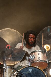 Drummer with dreadlocks focused on a drum kit with glittery drums and large cymbals on stage.