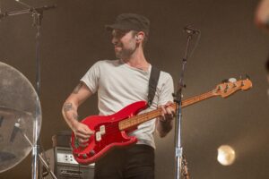 Male bass guitarist in a white t-shirt and black cap playing a red bass on stage.