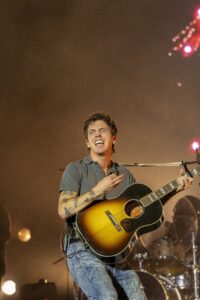Guitarist singing and playing a sunburst acoustic guitar on stage, stage lights glowing in the background, tattoos visible on his arm.