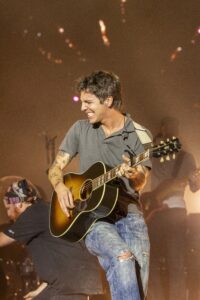 Male guitarist smiling while playing an acoustic guitar on stage during a concert, warm lights in the background.