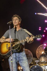 Male singer-songwriter playing an acoustic guitar on stage, singing into a microphone with drums and colorful lights in the background