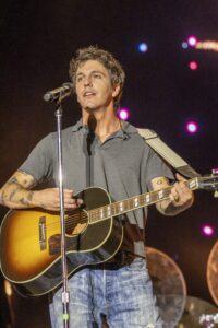 Male musician sings into a microphone while playing a sunburst acoustic guitar on stage with colorful lights.