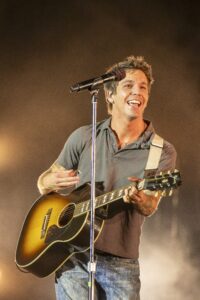 Male musician singing into a microphone while playing a sunburst acoustic guitar on stage, wearing a gray shirt and jeans.