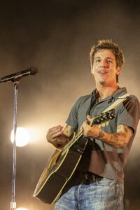 Male musician playing an acoustic guitar on stage, singing into a microphone with warm stage lights behind him.