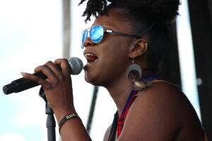 Female singer wearing blue mirrored sunglasses, singing into a microphone on stage.