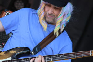 Person with rainbow-colored hair in a blue shirt plays a bass guitar, focusing on the neck and strings.