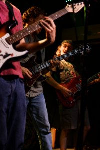 Three guitarists on stage playing electric guitars during a live band performance, with one smiling guitarist in the center and colorful stage lighting.