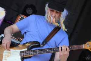 Musician with rainbow wig and blue shirt playing a sun-faded bass guitar on stage, focusing on frets and strings
