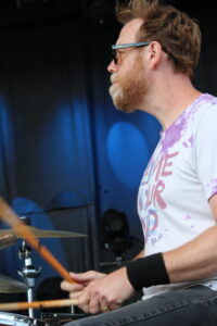 Side-profile of a bearded man wearing sunglasses, playing a drum kit with drumsticks on a stage with a blue backdrop