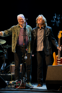 Two older musicians on a dimly lit stage, one in a floral shirt and headband, the other in a black jacket with arrows, with a guitar and gear nearby.