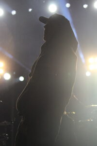 Silhouette of a performer wearing a hoodie and cap on a stage with bright backlighting from spotlights