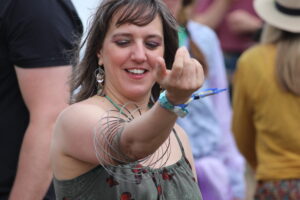 Smiling woman at an outdoor event extending her hand toward the camera, wearing a green dress with red floral print and bold jewelry.